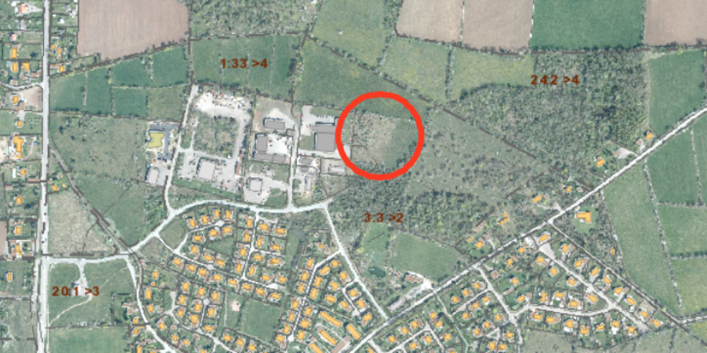 Flygfoto över ett bostadsområde och omgivande åkrar och skog, med ett planområde markerat med röd ring vid skogsbrynet nära industribyggnader. Synlig text: “1:33 >4”, “2:42 >4”, “3:3 >2”, “20:1 >3”, “2:6 >3”, “3:3 >4”, “3:3 >3”.