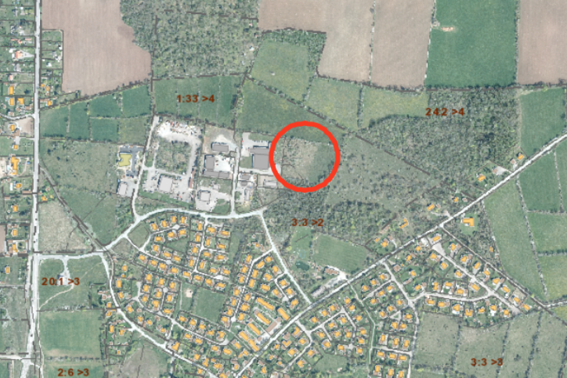 Flygfoto över ett bostadsområde och omgivande åkrar och skog, med ett planområde markerat med röd ring vid skogsbrynet nära industribyggnader. Synlig text: “1:33 >4”, “2:42 >4”, “3:3 >2”, “20:1 >3”, “2:6 >3”, “3:3 >4”, “3:3 >3”.
