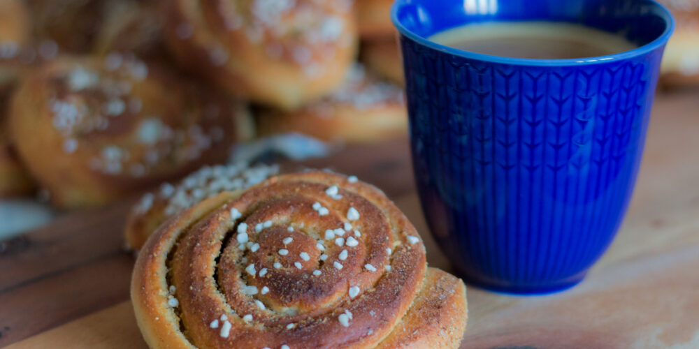 Blå kaffekopp fylld med kaffe står bredvid en kanelbulle med pärlsocker på en träbricka. I bakgrunden syns flera suddiga bullar, vilket skapar känslan av en fikapaus.