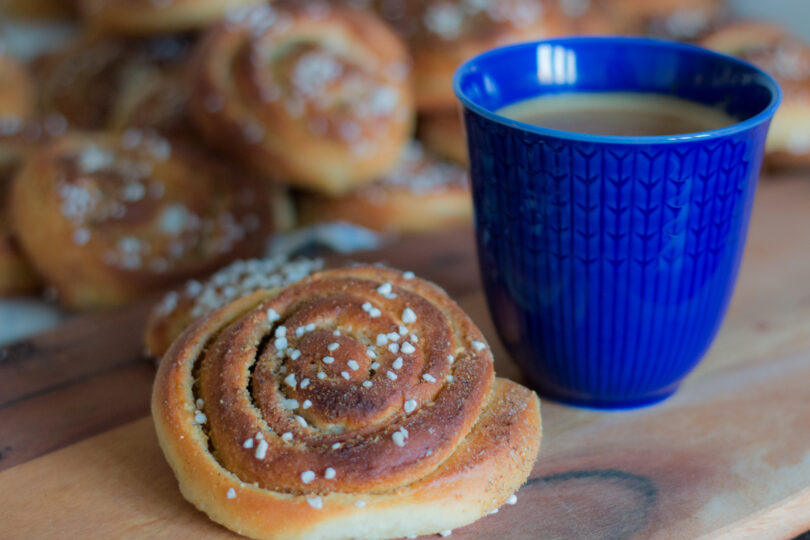 Blå kaffekopp fylld med kaffe står bredvid en kanelbulle med pärlsocker på en träbricka. I bakgrunden syns flera suddiga bullar, vilket skapar känslan av en fikapaus.