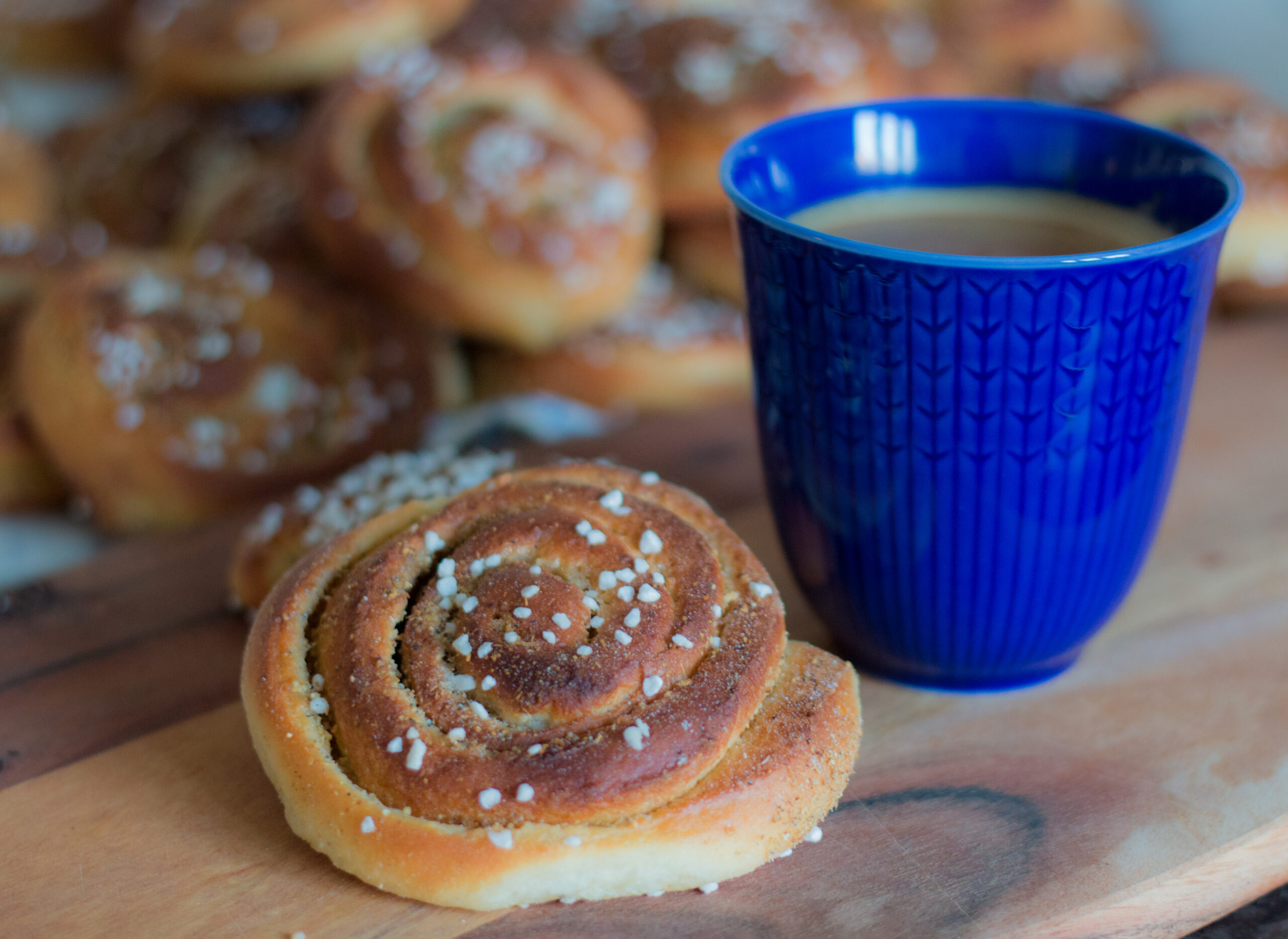 Blå kaffekopp fylld med kaffe står bredvid en kanelbulle med pärlsocker på en träbricka. I bakgrunden syns flera suddiga bullar, vilket skapar känslan av en fikapaus.