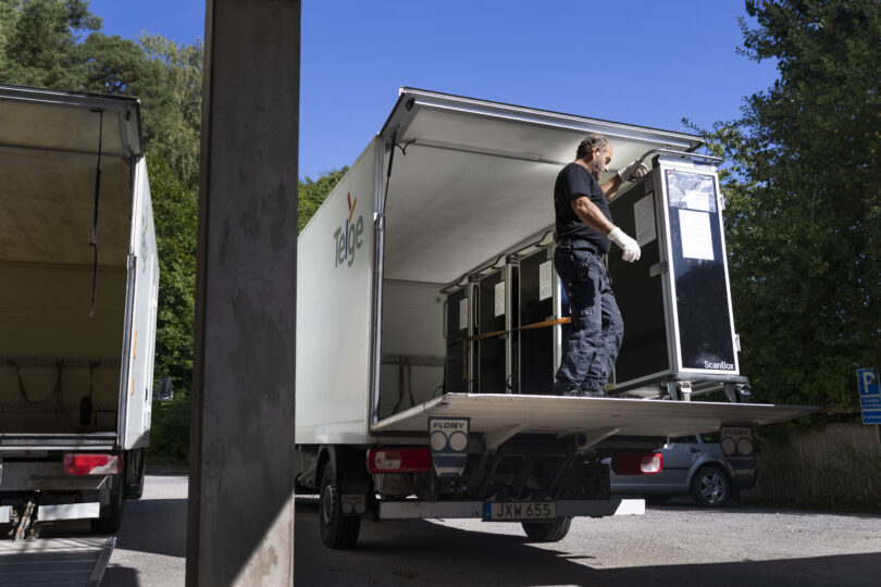 Man i arbetskläder står på bakgavellyften och skjuter en stor svart rullvagn märkt ”ScanBox” in i en lastbil med öppna dörrar och texten ”Tagde”, parkerad utomhus bland träd i solsken.