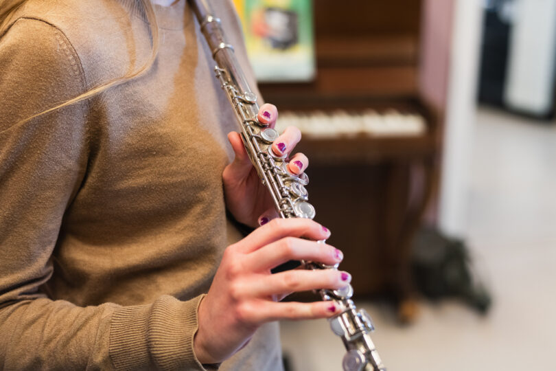 Barns händer med lila nagellack håller och spelar en tvärflöjt, med fingrarna på klaffarna. Närbild i inomhusmiljö med suddig bakgrund, som antyder en musiklektion i kulturskola.