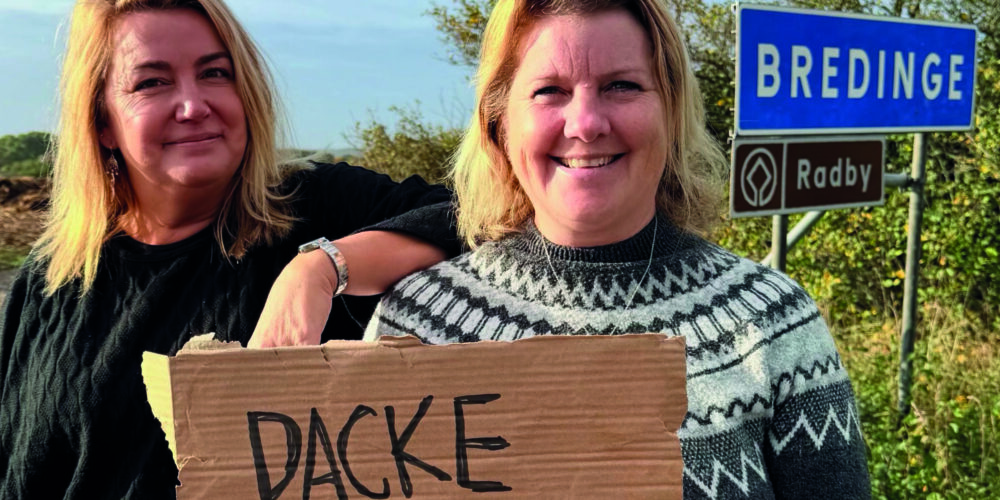 Två kvinnor ler mot kameran vid en vägkant; den ena håller en kartongskylt med texten ”BACKE WAS HERE”. I bakgrunden syns vägskyltarna ”BREDINGE” och ”Radby” samt rubriken ”KALKSTENS KANALEN”.