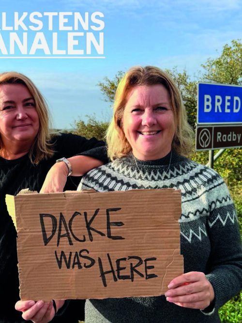 Två kvinnor ler mot kameran vid en vägkant; den ena håller en kartongskylt med texten ”BACKE WAS HERE”. I bakgrunden syns vägskyltarna ”BREDINGE” och ”Radby” samt rubriken ”KALKSTENS KANALEN”.