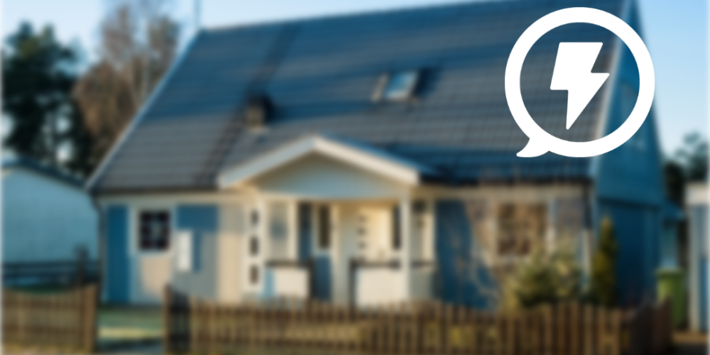 Villa med mörkt tak och solpaneler ligger i fokus bakom ett staket, fotograferad i dagsljus. Uppe till höger syns en vit pratbubbla med en blixtsymbol, som antyder el eller energiförbrukning.