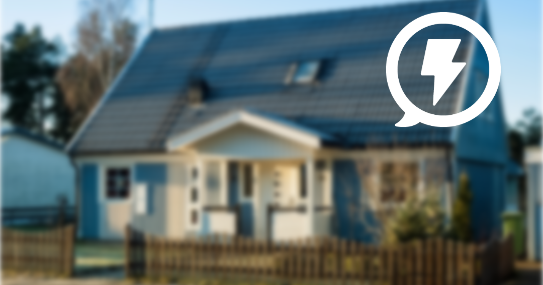 Villa med mörkt tak och solpaneler ligger i fokus bakom ett staket, fotograferad i dagsljus. Uppe till höger syns en vit pratbubbla med en blixtsymbol, som antyder el eller energiförbrukning.