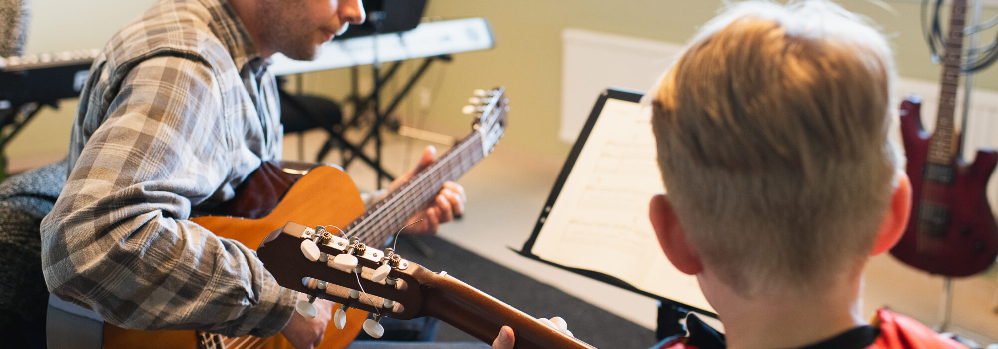Gitarrlärare sitter mittemot en elev och spelar akustisk gitarr medan eleven håller sin egen gitarr och tittar mot ett notställ med noter. Musikrums­miljö med keyboard i bakgrunden och elgitarr bredvid fönstret.