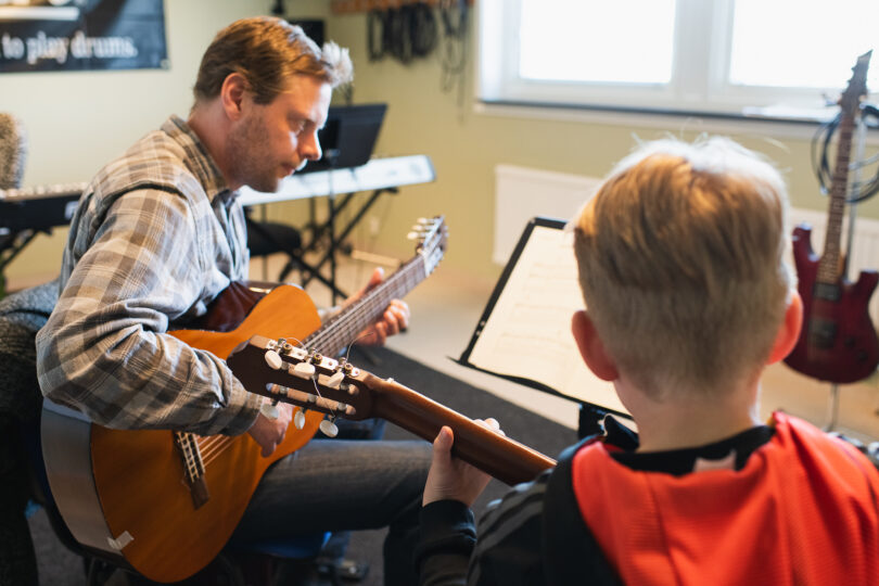 Gitarrlärare sitter mittemot en elev och spelar akustisk gitarr medan eleven håller sin egen gitarr och tittar mot ett notställ med noter. Musikrums­miljö med keyboard i bakgrunden och elgitarr bredvid fönstret.