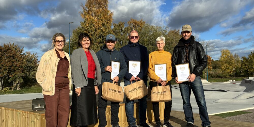 Sex personer poserar på en träplattform utomhus och håller inramade diplom samt papperskassar med texten ”MYSINGE KÖTT”. Bakom dem syns höstträd och en dramatisk molnhimmel över en park/skatepark.