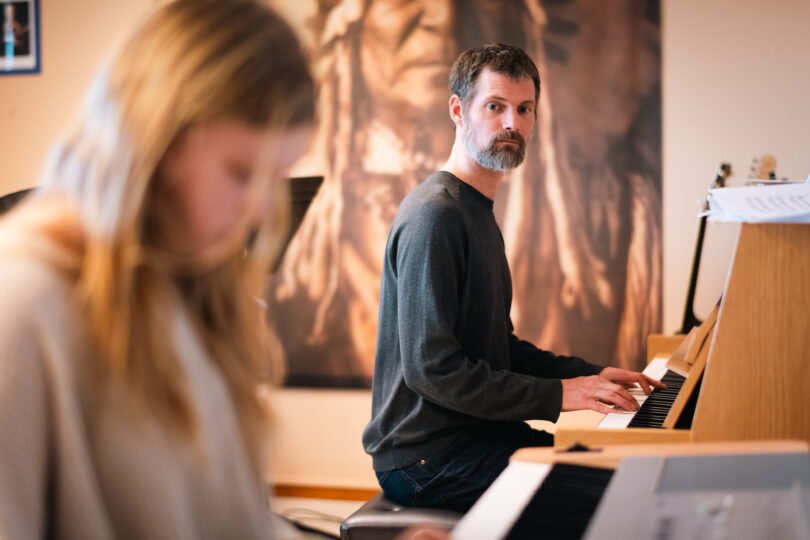 Skäggig man sitter vid ett upprätt piano och spelar med båda händerna, medan han tittar åt sidan. I förgrunden syns en suddig elev; i bakgrunden finns en stor väggbild i ett musikklassrum.