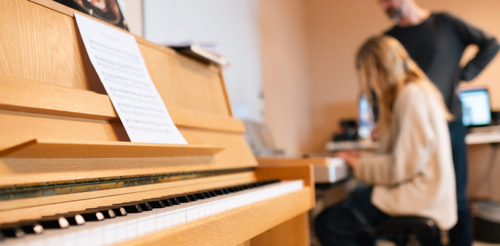 Notblad på ett ljust upprätt piano står i fokus, medan en elev sitter vid ett annat piano och spelar. En lärare står bredvid och lyssnar i ett klassrum, med dator på bordet.