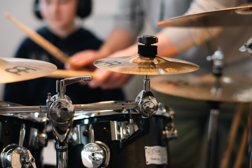 Närbild på cymbaler och stativ på ett trumset medan en elev i bakgrunden spelar med trumpinnar. Fokus ligger på blänkande metall och cymbalens yta, med suddig musiklektion som miljö.