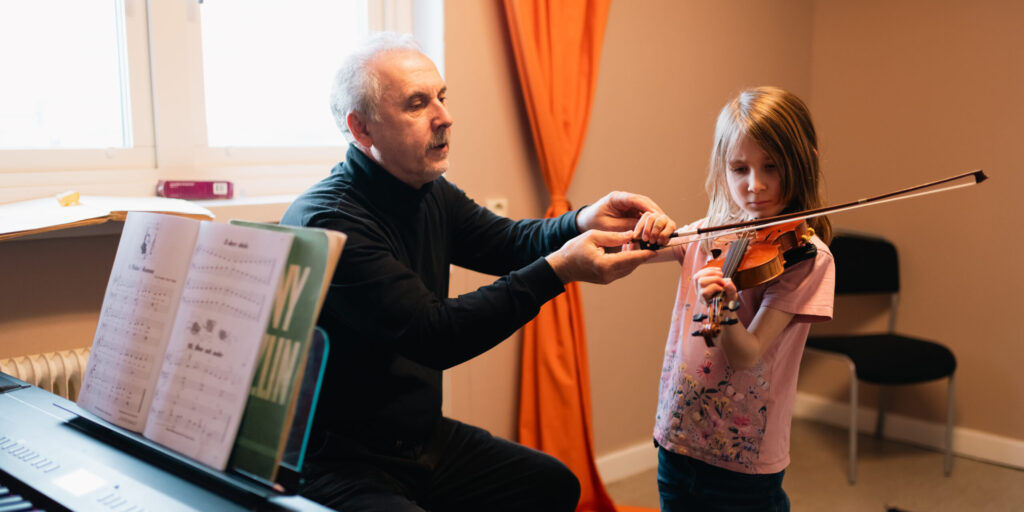 Violinlärare justerar en flickas stråkhand medan hon håller fiolen under hakan och övar. Lektionen sker i ett klassrum med orange gardin; i förgrunden syns ett keyboard med notställ och noter.