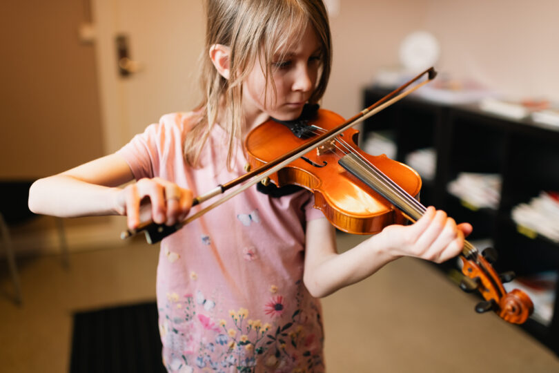 Flicka spelar violin med koncentrerad blick, håller instrumentet mot axeln och drar stråken över strängarna. Inomhusmiljö med suddig bakgrund och varm belysning, hon bär rosa tröja med blommönster.
