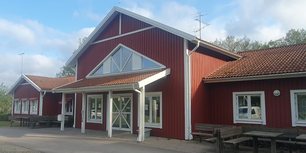 Röd träbyggnad med vitmålade detaljer och tegeltak står i soligt väder. Entrén har glasdörrar under ett utskjutande tak, och flera fönster syns. Utanför finns träbänkar och bord, med gröna träd i bakgrunden.