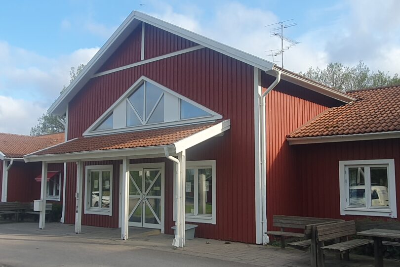 Röd träbyggnad med vitmålade detaljer och tegeltak står i soligt väder. Entrén har glasdörrar under ett utskjutande tak, och flera fönster syns. Utanför finns träbänkar och bord, med gröna träd i bakgrunden.