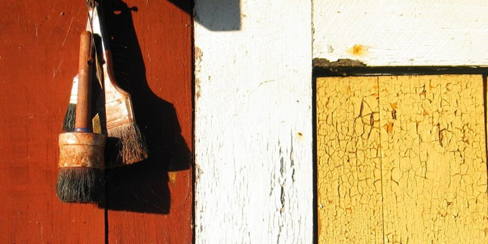 Två slitna penslar hänger på en spik mot en röd trävägg, belysta av starkt solljus. Intill syns vit panel och gul, krackelerad färg som framhäver väderbitet trä och struktur.