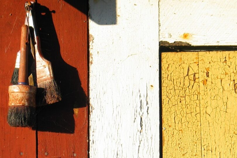 Två slitna penslar hänger på en spik mot en röd trävägg, belysta av starkt solljus. Intill syns vit panel och gul, krackelerad färg som framhäver väderbitet trä och struktur.