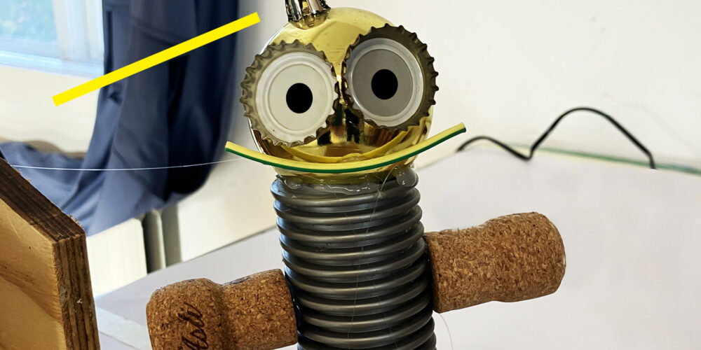Hemmagjord skrotrobot står upprätt på ett bord, med korkarmar, ribbad grå kropp och guldfärgat huvud med stora ögon. Gula strålar markerar huvudet. Text längst ned: “SKROTBOTAR”.