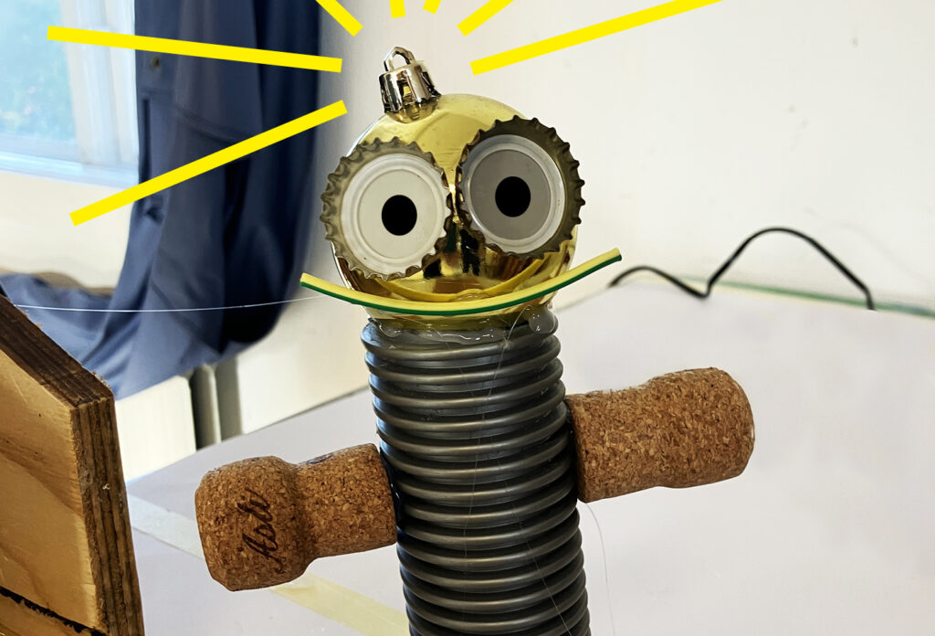 Hemmagjord skrotrobot står upprätt på ett bord, med korkarmar, ribbad grå kropp och guldfärgat huvud med stora ögon. Gula strålar markerar huvudet. Text längst ned: “SKROTBOTAR”.