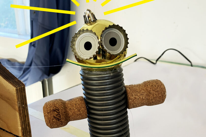 Hemmagjord skrotrobot står upprätt på ett bord, med korkarmar, ribbad grå kropp och guldfärgat huvud med stora ögon. Gula strålar markerar huvudet. Text längst ned: “SKROTBOTAR”.